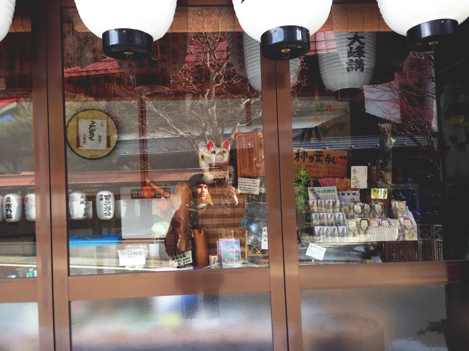 A shopfront window in a pilgrimage town hung with paper lanterns, a cat mask and local goods inside, and Lindsey's reflection in the glass as she takes the photograph.