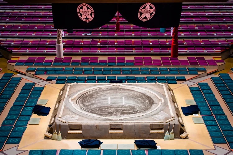 An empty sumo arena with the raised clay dohyō ring at center, ringed by cushioned seating, brooms resting at its base.