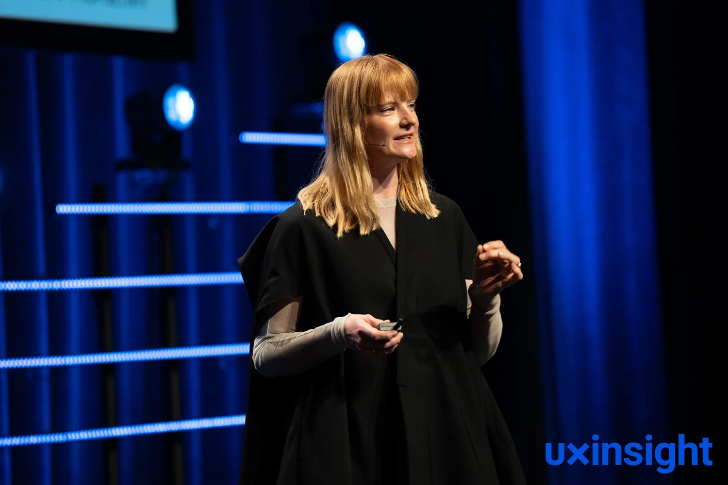 Lindsey delivering a keynote on a blue-lit conference stage, wearing a headset microphone and gesturing mid-sentence.