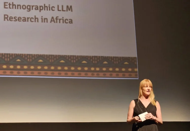 Lindsey on stage in a black dress beneath a projected slide titled Ethnographic LLM Research in Africa.