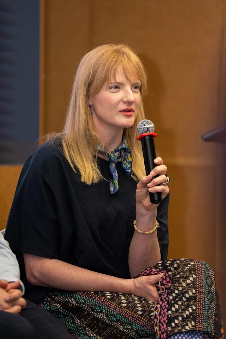 Lindsey seated on a panel, speaking into a handheld microphone, wearing a dark top with a floral scarf.