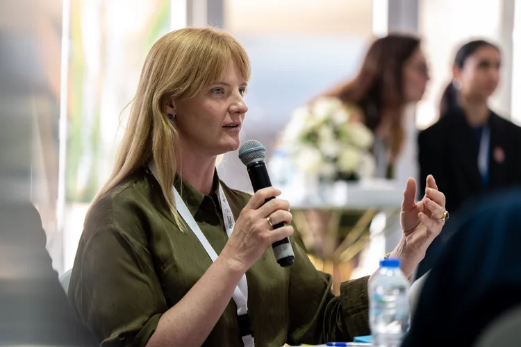 Lindsey in an olive shirt speaking into a handheld microphone at a roundtable, with flowers and other participants blurred behind.