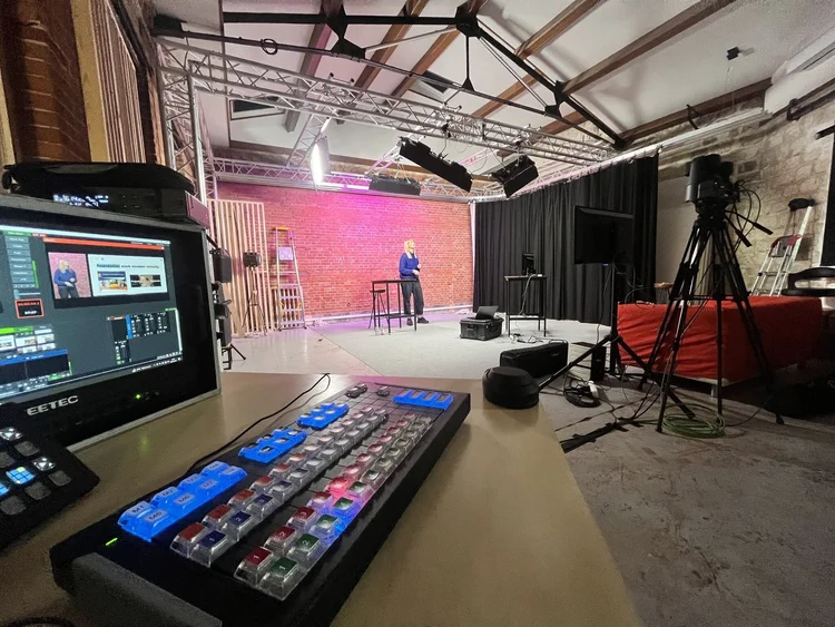 View from a video control desk of Lindsey presenting at a lectern against a brick wall, surrounded by studio cameras and truss lighting.