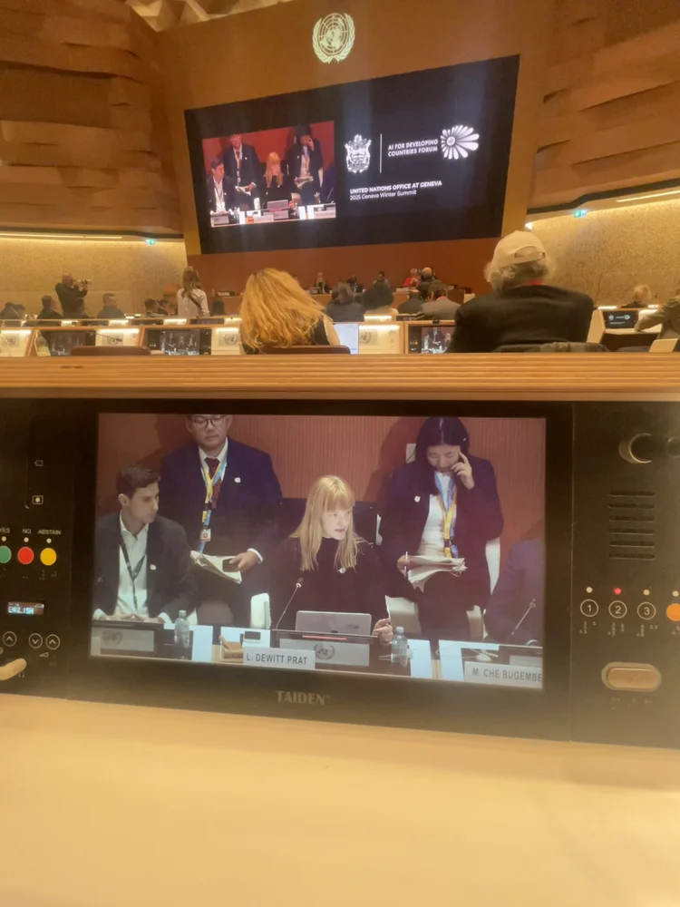 Conference hall at the United Nations Office at Geneva during the AI for Developing Countries Forum, with a desk monitor showing Lindsey speaking behind a nameplate reading L. DeWitt Prat.