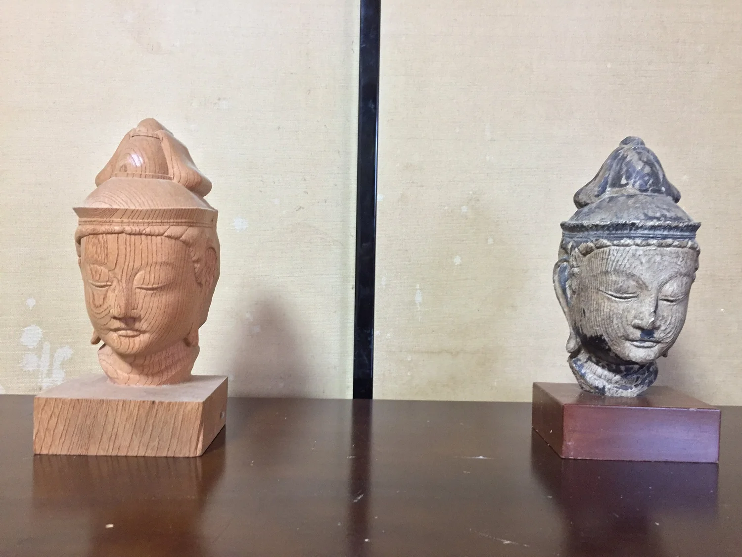 Two carved bodhisattva heads on wooden bases, one in pale fresh-cut wood and one aged and darkened.