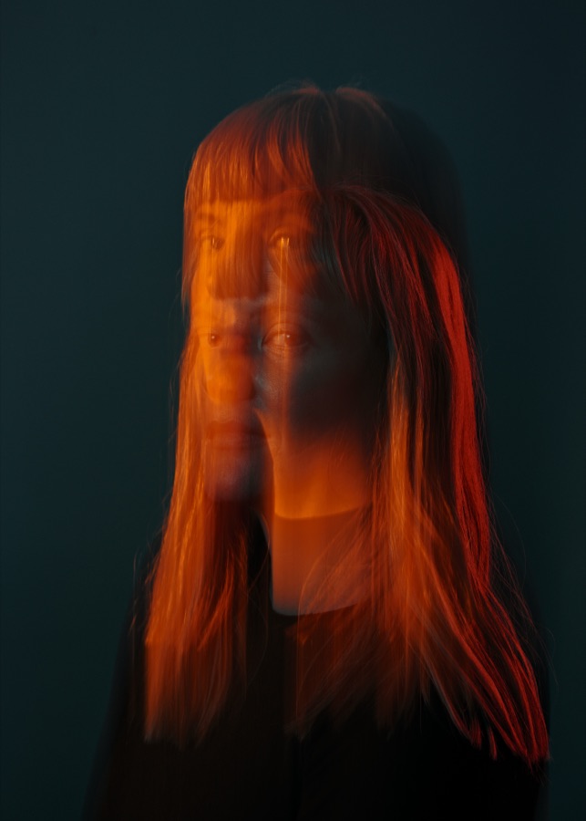 Portrait of Lindsey DeWitt Prat in a long exposure, her face dissolved behind streaks of orange light and hair against deep teal. Photo by Jan Opdekamp.