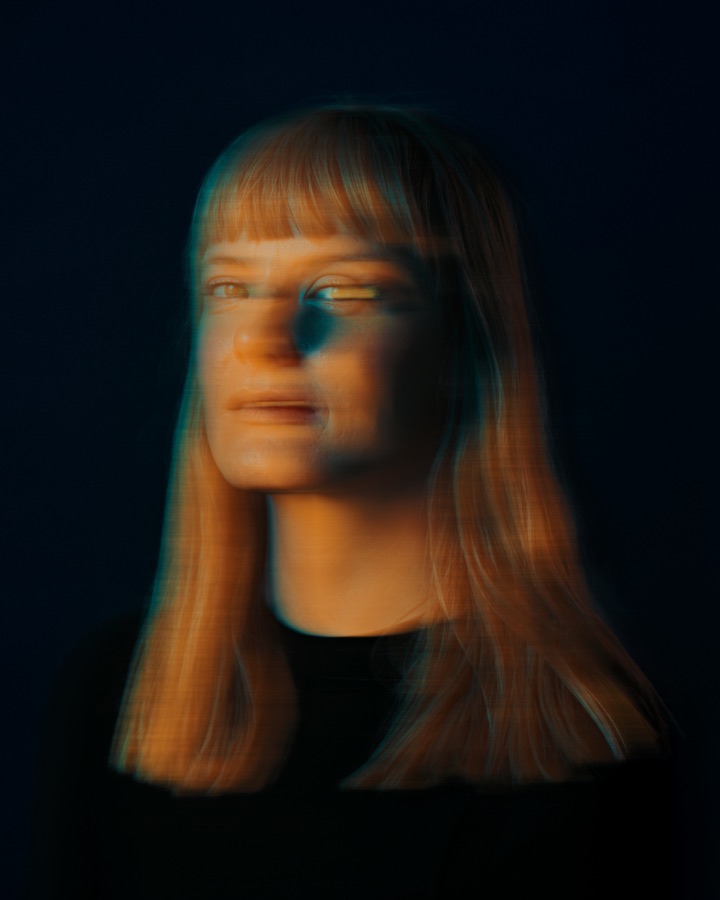 Portrait of Lindsey DeWitt Prat facing the camera, her face softly doubled by a long exposure, lit in orange and teal against a dark ground. Photo by Jan Opdekamp.