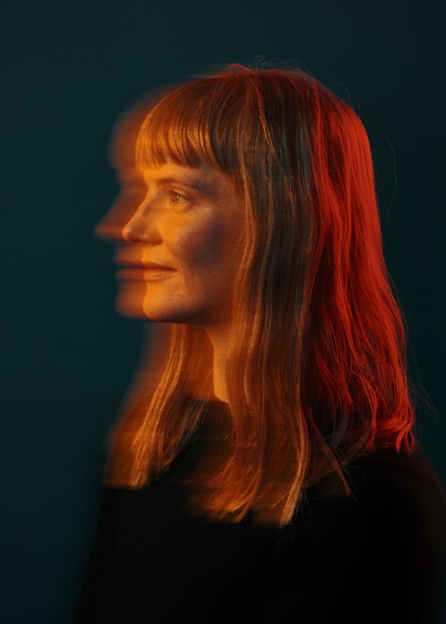Profile portrait of Lindsey DeWitt Prat in warm orange light, a second exposure of her profile trailing behind the first against deep teal. Photo by Jan Opdekamp.