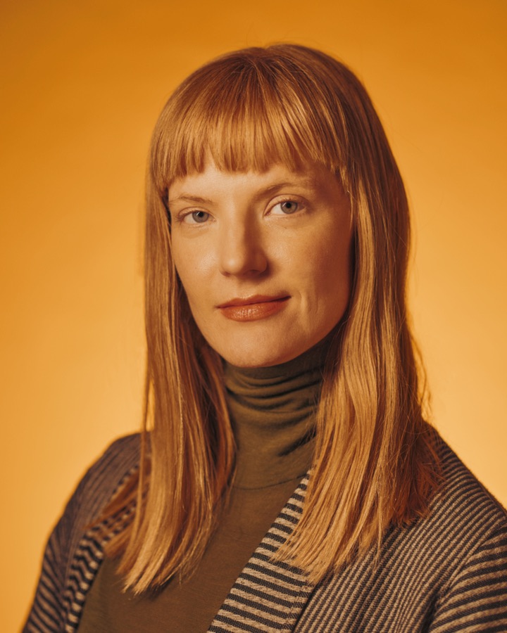 Portrait of Lindsey DeWitt Prat looking into the camera against a warm amber background, wearing an olive turtleneck and striped jacket. Photo by Jan Opdekamp.